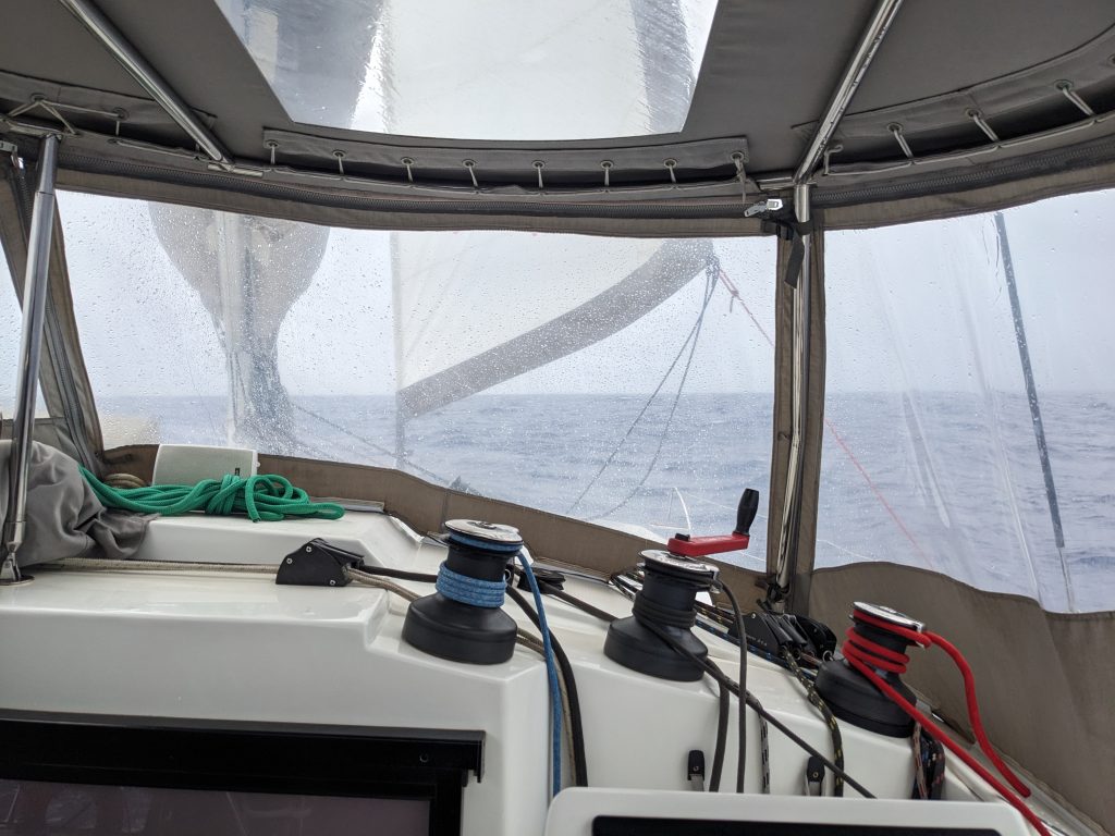 Image of the cockpit of a sailboat, with the winches and sail control lines visible, view of the sea out in front blocked by the cock[it covering, on which we can see raindrops.