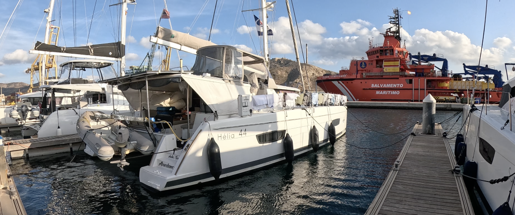 Image of sailing catamaran in its marina slip, with red commercial vessel in the background