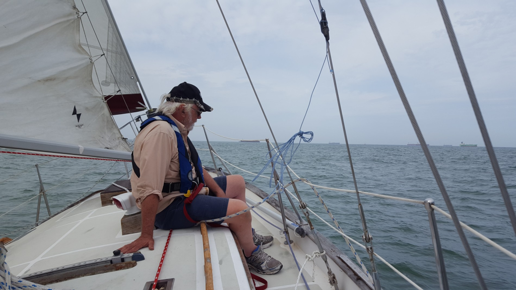 Image of grey-haired man seated near the bow of a sailboat underway