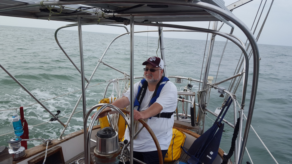 Image of grey-bearded sailor at the helm of a sailboat under way