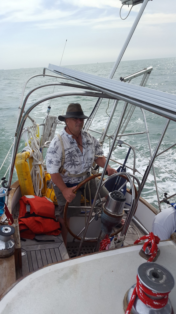 Image of barrel-chested man at the helm of a sailboat under way